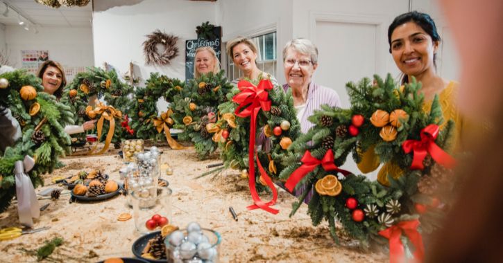 group of women smiling at camera and holding up Christmas wreaths they have made.